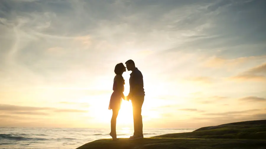 A couple in silhouette kisses on a rock overlooking a sunset by the ocean